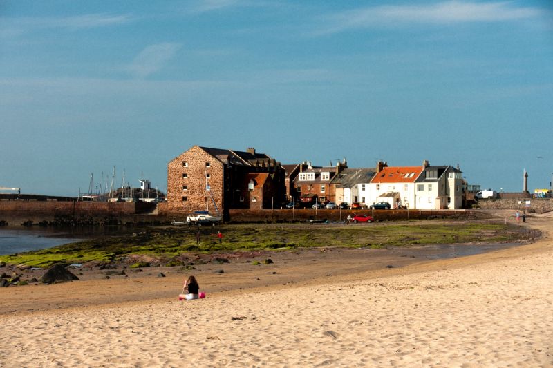 A sandy beach with one person sitting on the shore, colorful houses and brick buildings in the background, and a bright blue sky overhead—an ideal spot for students seeking relaxing day trips from Edinburgh.