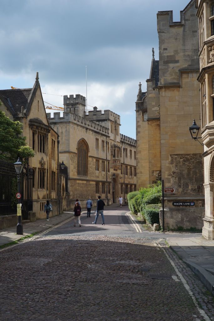 A cobblestone street lined with historic stone buildings, a few people—including some students—walking, and a partly cloudy sky above. Signs on the right indicate “Magpie Lane,” suggesting a picturesque spot perfect for day trips from Coventry.