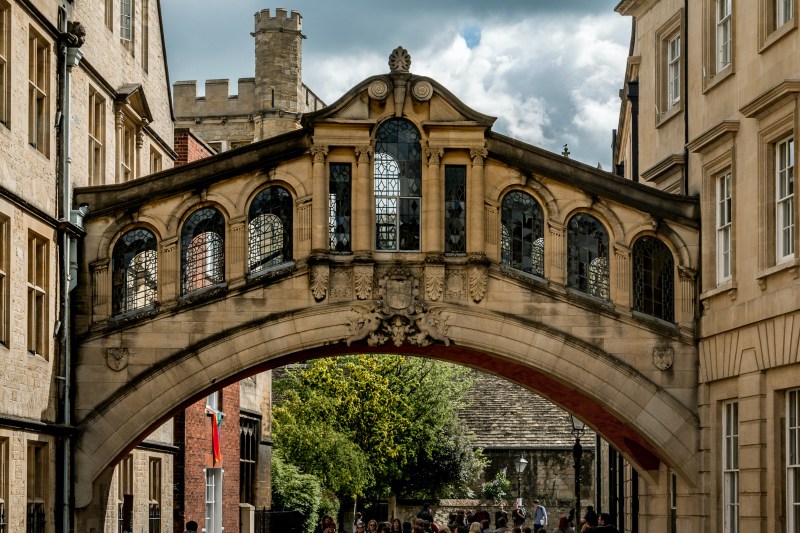 A stone arched bridge with ornate windows connects two historic buildings above a narrow street in Coventry, with students and other people walking underneath and a tower in the background—a perfect scene for memorable day trips.