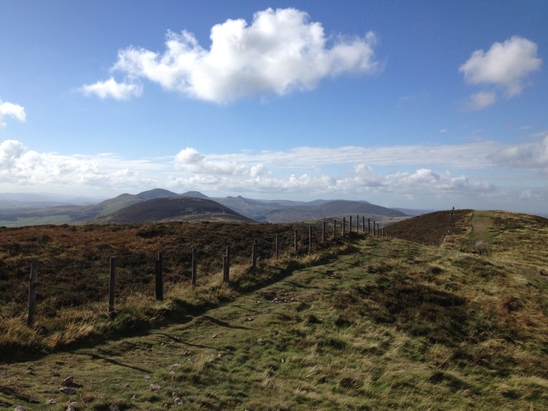 A grassy hilltop with a wooden fence running along the ridge, popular with students on day trips from Edinburgh, overlooks distant mountains under a blue sky with scattered clouds.