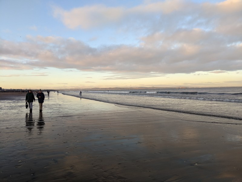 People, including students on day trips from Edinburgh, walk along a wet, reflective beach at sunset, with gentle waves and a cloudy sky in the background.