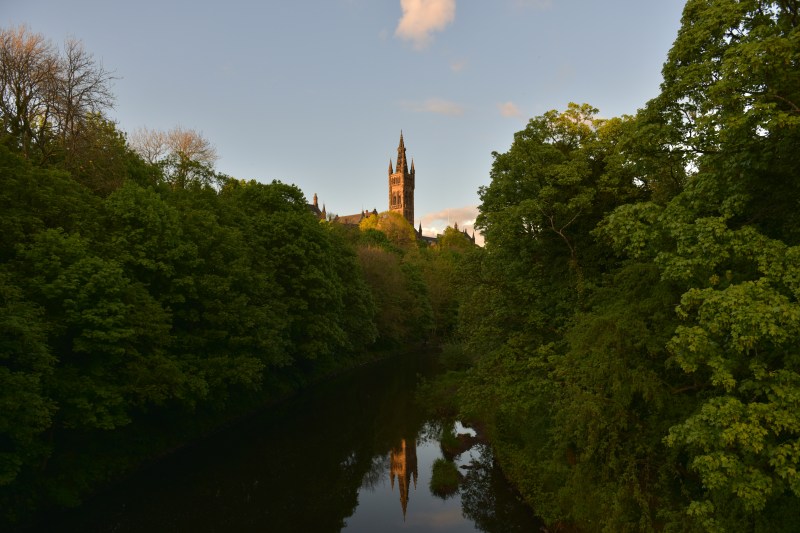 A tall, gothic-style tower rises above dense, green trees at sunset in the West End, its reflection visible in the calm river below. The sky is partly cloudy with warm sunlight illuminating parts of the scene, making it a picturesque spot popular among students looking for memorable things to do.