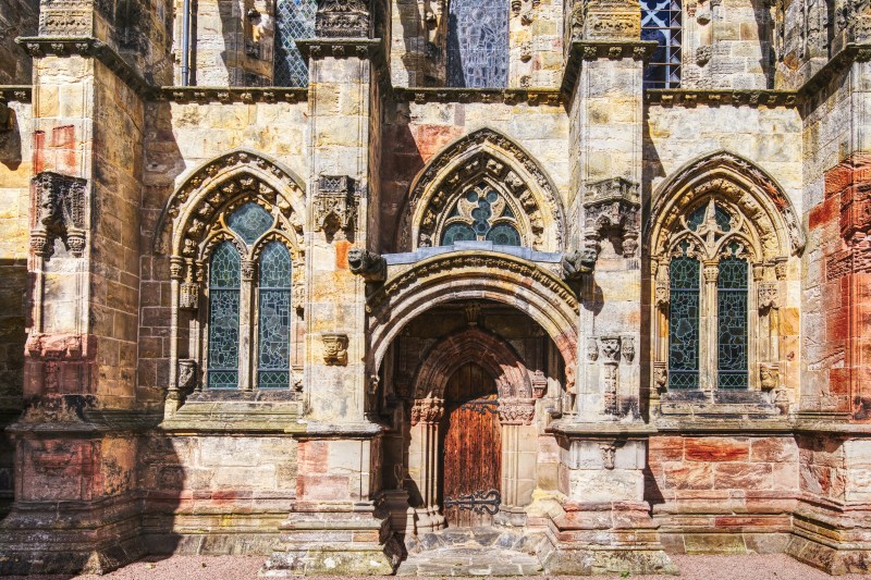 Stone gothic church facade with arched wooden door and three pointed stained-glass windows, featuring ornate carvings and weathered textures in warm yellow, brown, and red tones, seen in bright daylight—a striking landmark often admired by students on day trips exploring the historic heart of Edinburgh.