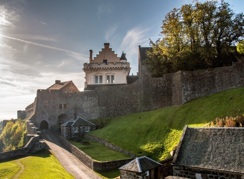 A historic stone fortress with tall walls and a building featuring a stepped gable roof—perfect for students seeking memorable day trips from Edinburgh—surrounded by green grass and trees, under a partly cloudy sky.