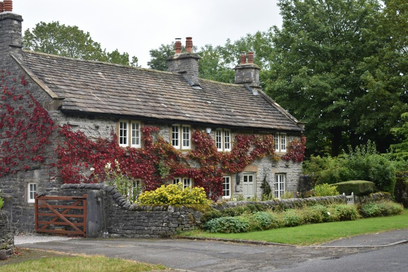 A stone cottage with a tiled roof and climbing red ivy, surrounded by a stone wall, a wooden gate, and a garden, perfect for students on Birmingham day trips.