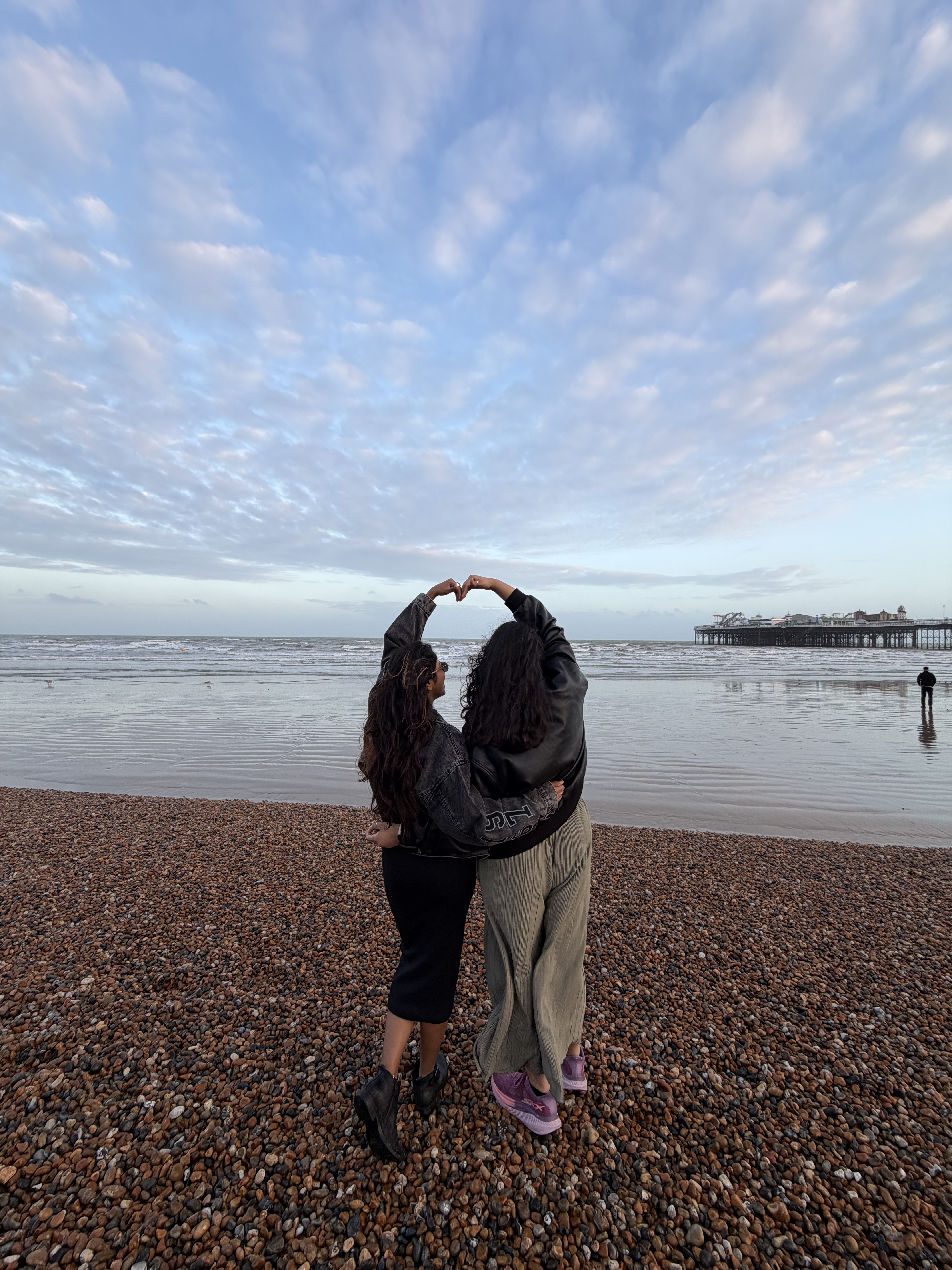 Two people stand on a pebbled beach facing the sea, arms raised together to form a heart shape—capturing a Vita moment of forever friends under a cloudy sky, with a pier stretching into the water behind them.