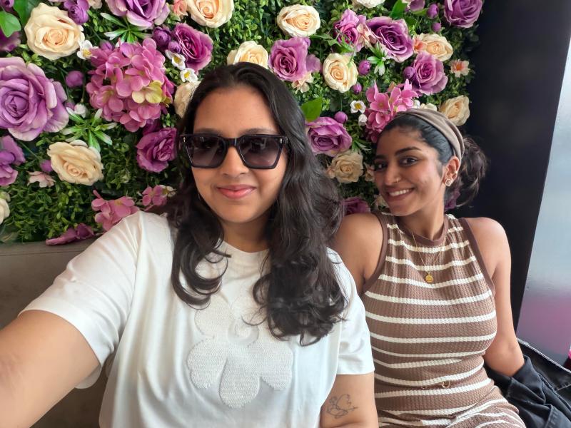 Two women smiling—one in sunglasses and a white shirt, the other in a striped top—sit before a wall of purple, pink, and white flowers, capturing a Vita moment of Forever Friends.