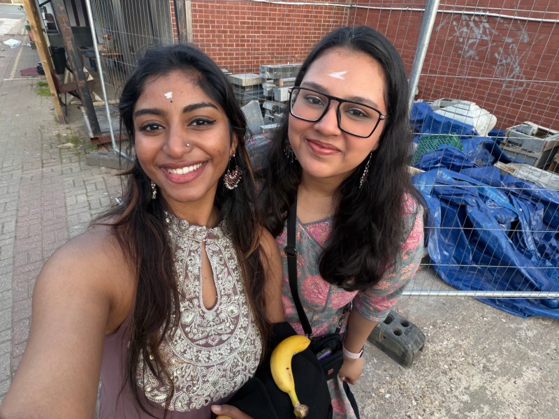 Two smiling South Asian women in traditional attire pose for a selfie outdoors, capturing their forever friends moment—one in metallic embroidery, the other in glasses and a floral outfit with a banana.