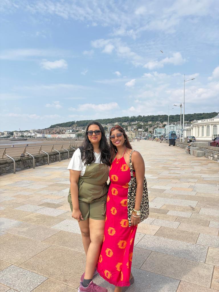 Two women smile on a seaside promenade—one in green overalls and sunglasses, the other in a red dress with a leopard-print bag. Their joyful pose captures true Forever Friends moments by the beach under a partly cloudy sky.