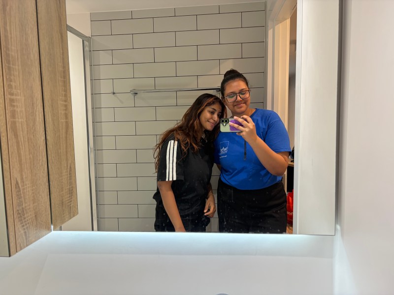 Two people stand together in front of a bathroom mirror, smiling. Sharing their Student Stories, one wears stripes while the other, in blue with glasses, snaps the photo—capturing the spirit of Forever Friends.