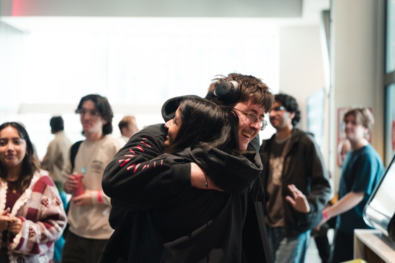 Two people wearing black hoodies share an embrace and smile, reflecting a successful partnership in a room with others in the background.
