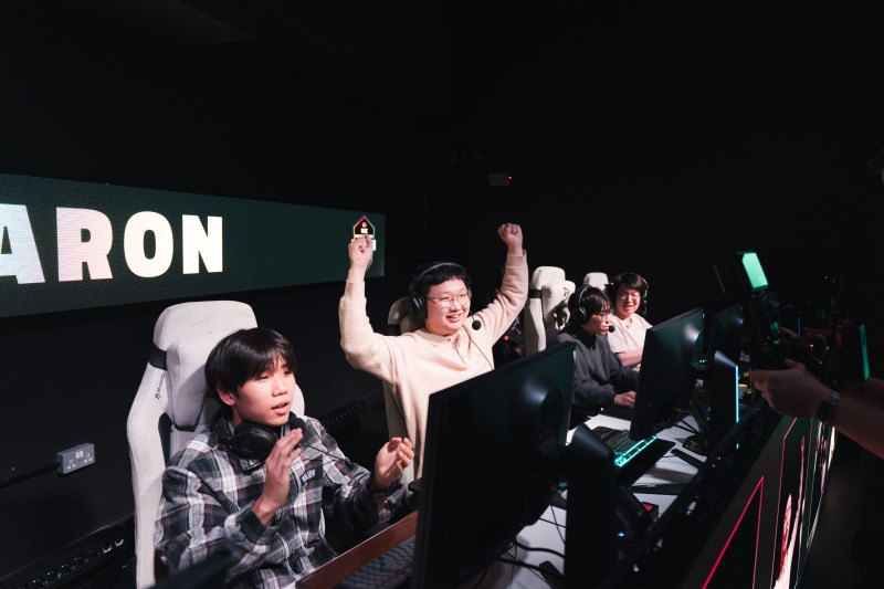 Four people playing video games at computers in a display of collaboration, with one player cheering with arms raised in victory.