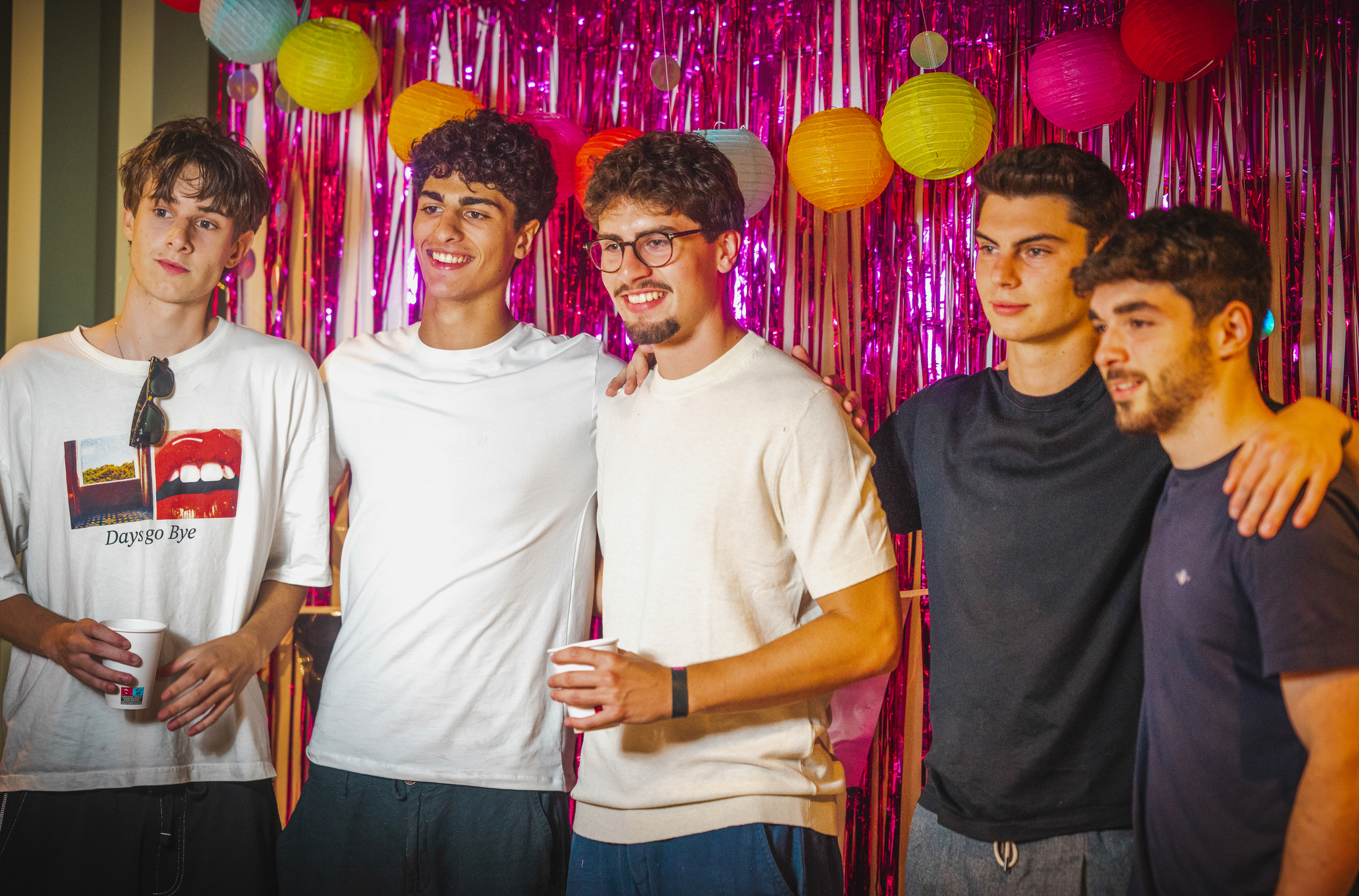 Five young men standing together in front of a pink tinsel backdrop decorated with paper lanterns.