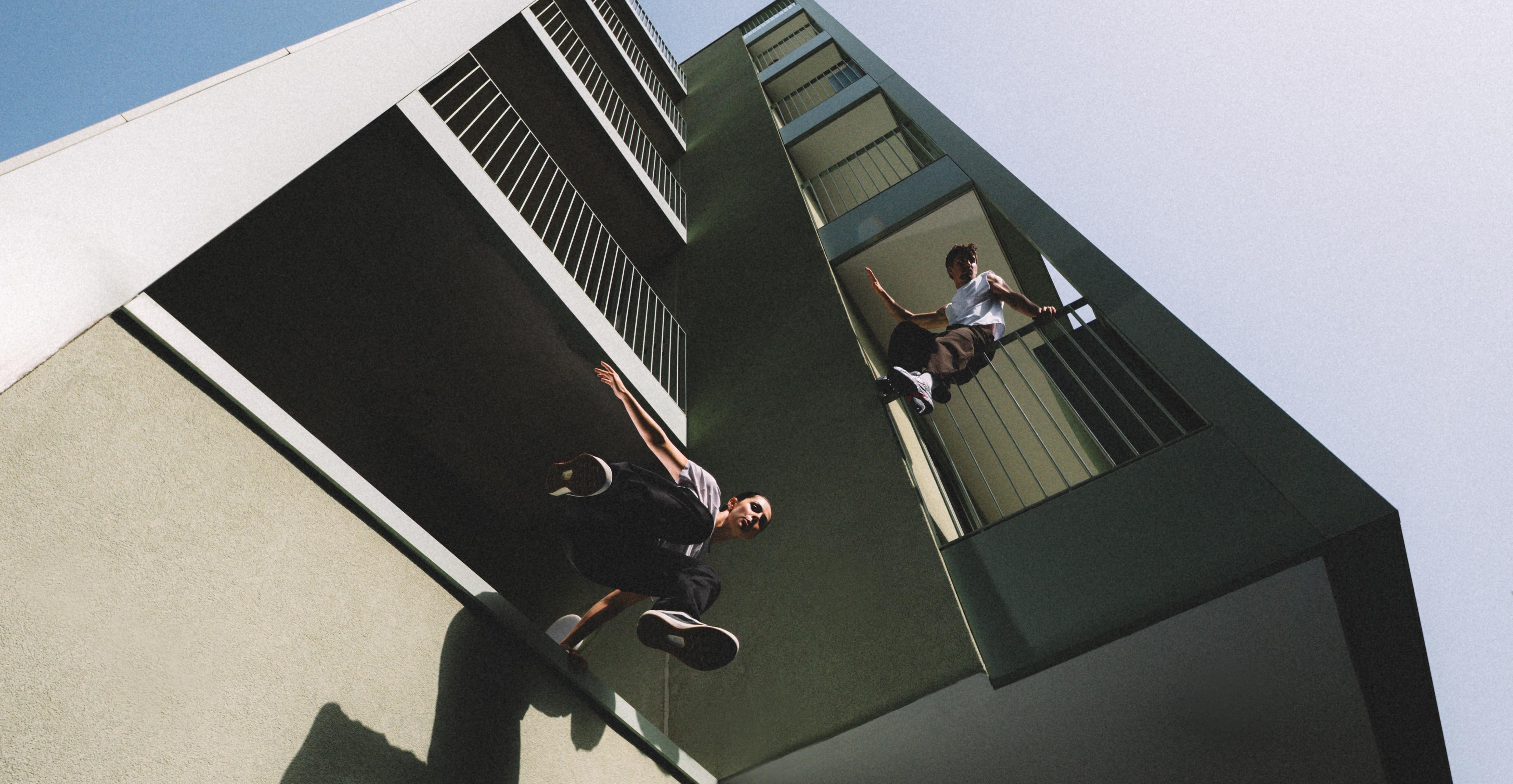 Low-angle view of two men on a building exterior, with one perched on a balcony railing and another leaping from a ledge.