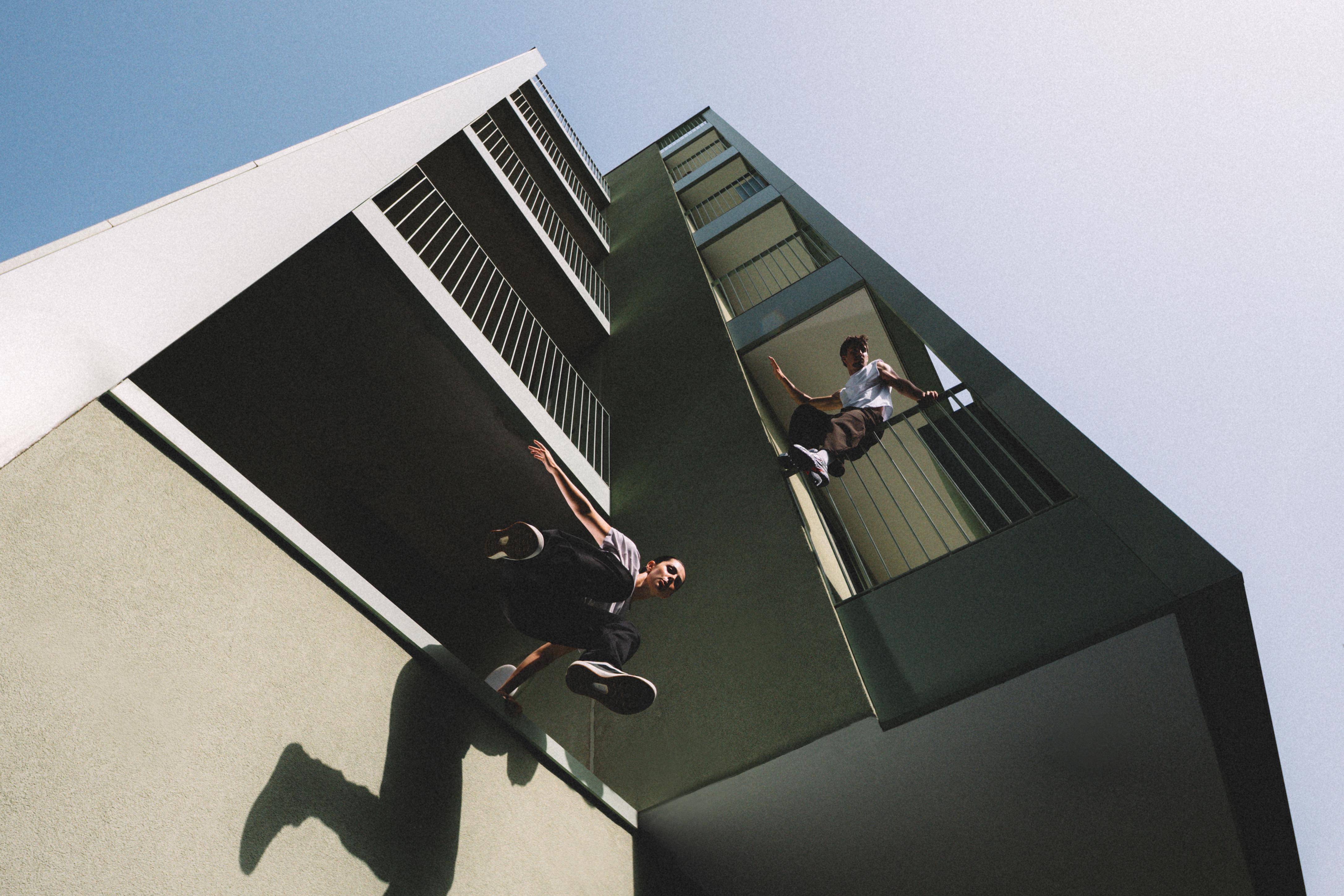 Low-angle view of two men on a building exterior, with one perched on a balcony railing and another leaping from a ledge.