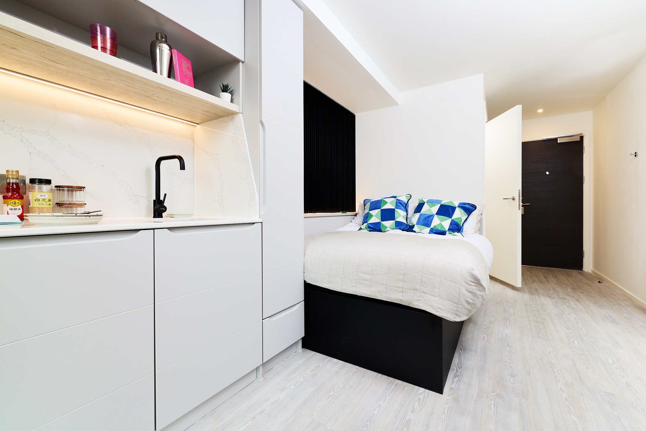 Studio apartment featuring a kitchen with white cabinetry and a black faucet, beside a bed with blue and green patterned pillows.