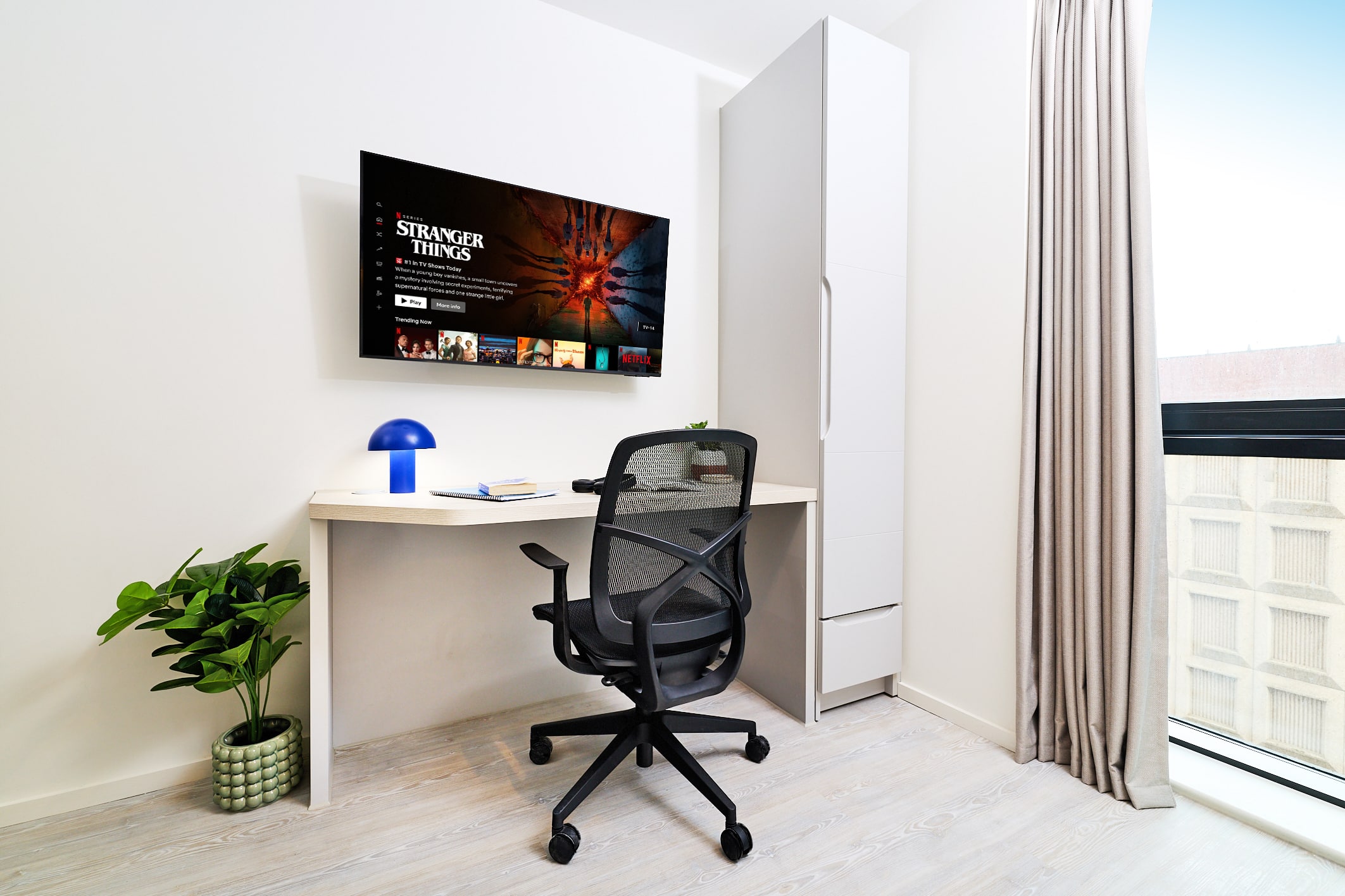 A home office nook with a wall-mounted television displaying Netflix, a desk with a blue lamp, a black mesh office chair, and a tall white cabinet.
