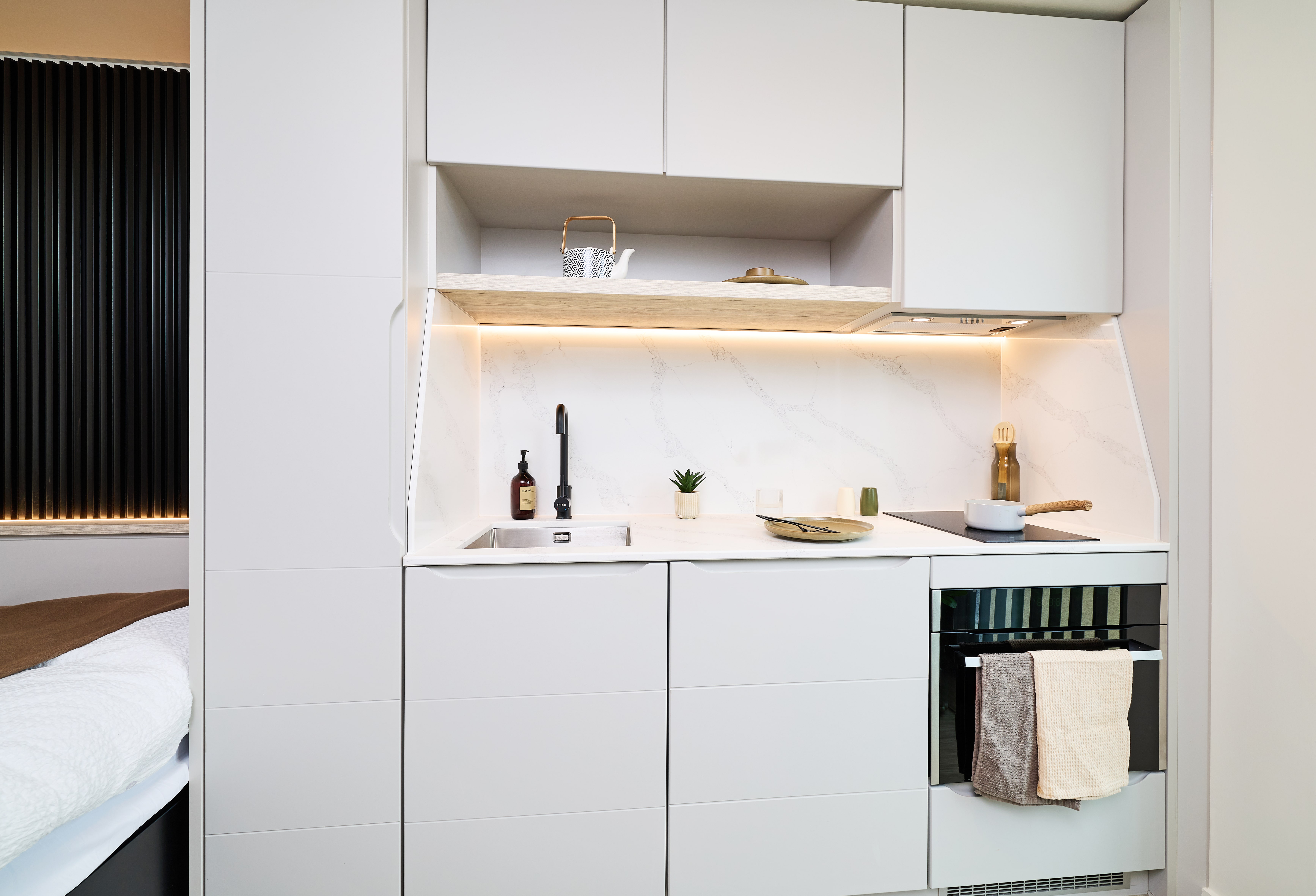 A modern kitchen unit with white cabinets, a sink, a stovetop, and a built-in oven, featuring a marble-style backsplash and lit shelving.
