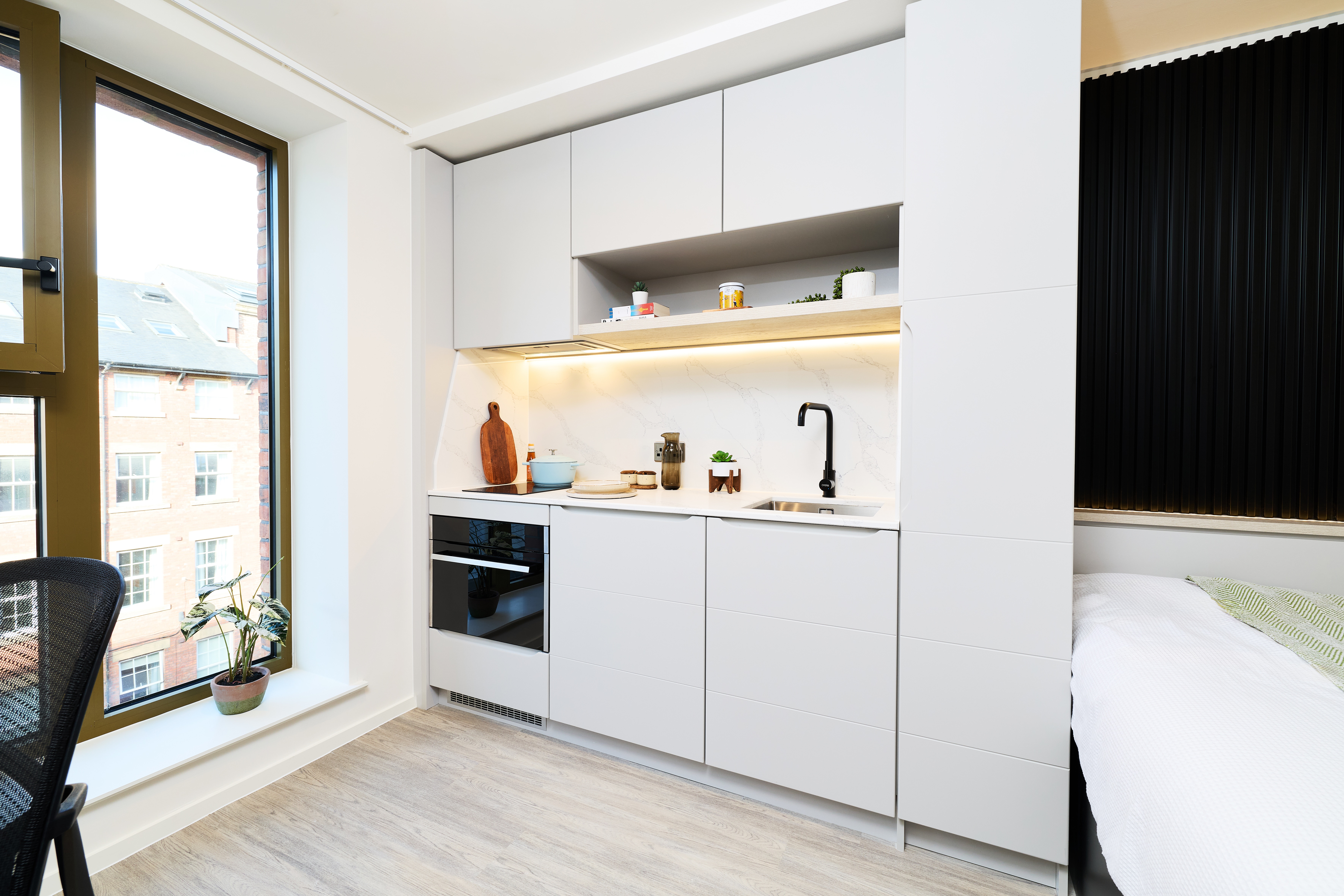 A compact kitchen unit with light gray cabinets, a built-in oven, marble-patterned backsplash, and integrated sink, situated next to a window and a bed.