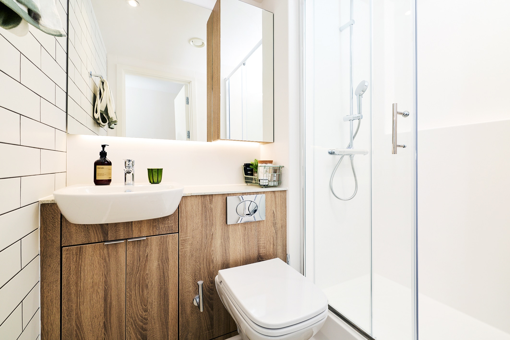 A modern bathroom featuring a wall-mounted sink, a wood-grain vanity cabinet, a toilet, and a glass-enclosed shower.