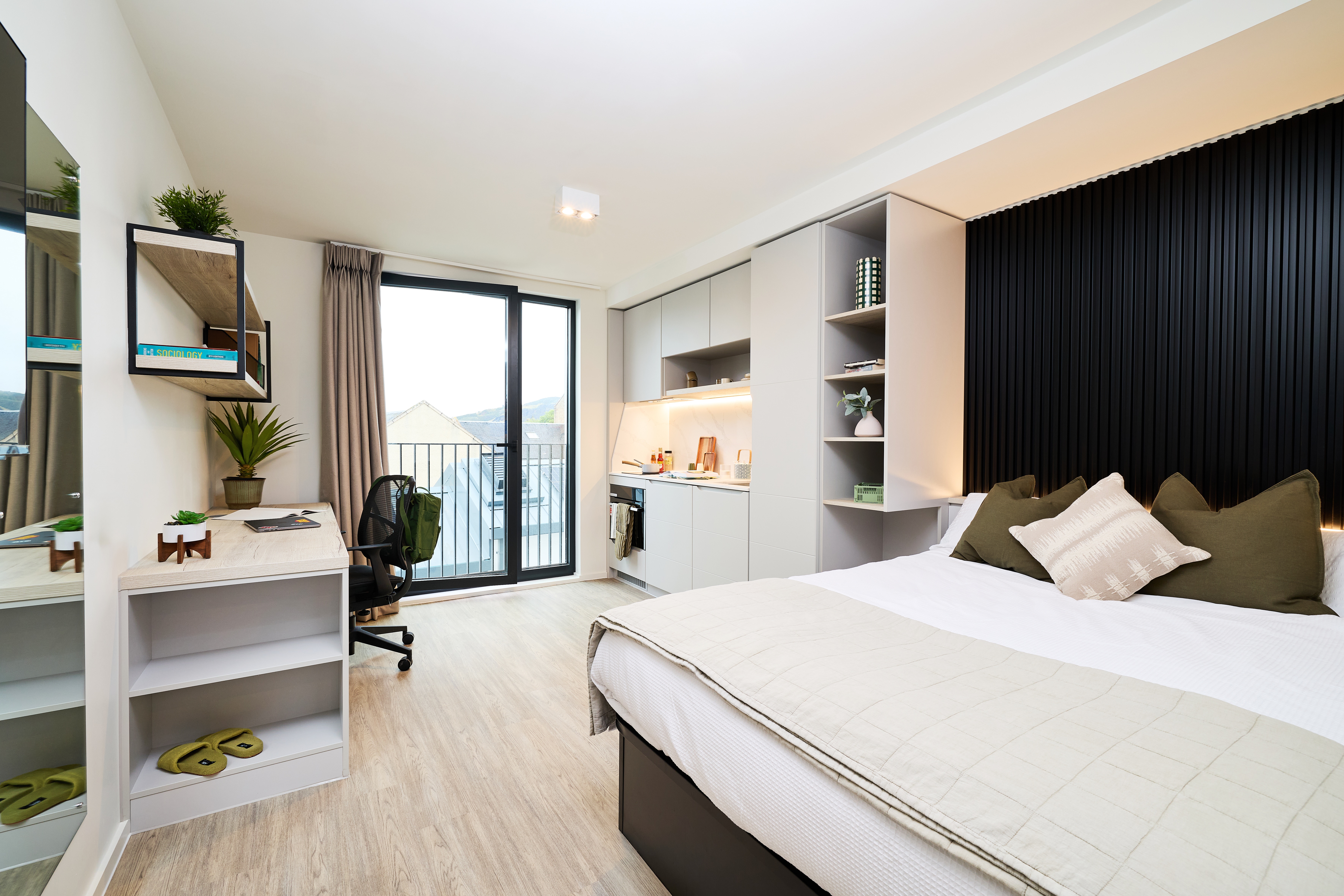 A studio apartment featuring a bed with olive green pillows, a desk with a computer chair, and a compact kitchenette with integrated shelving.
