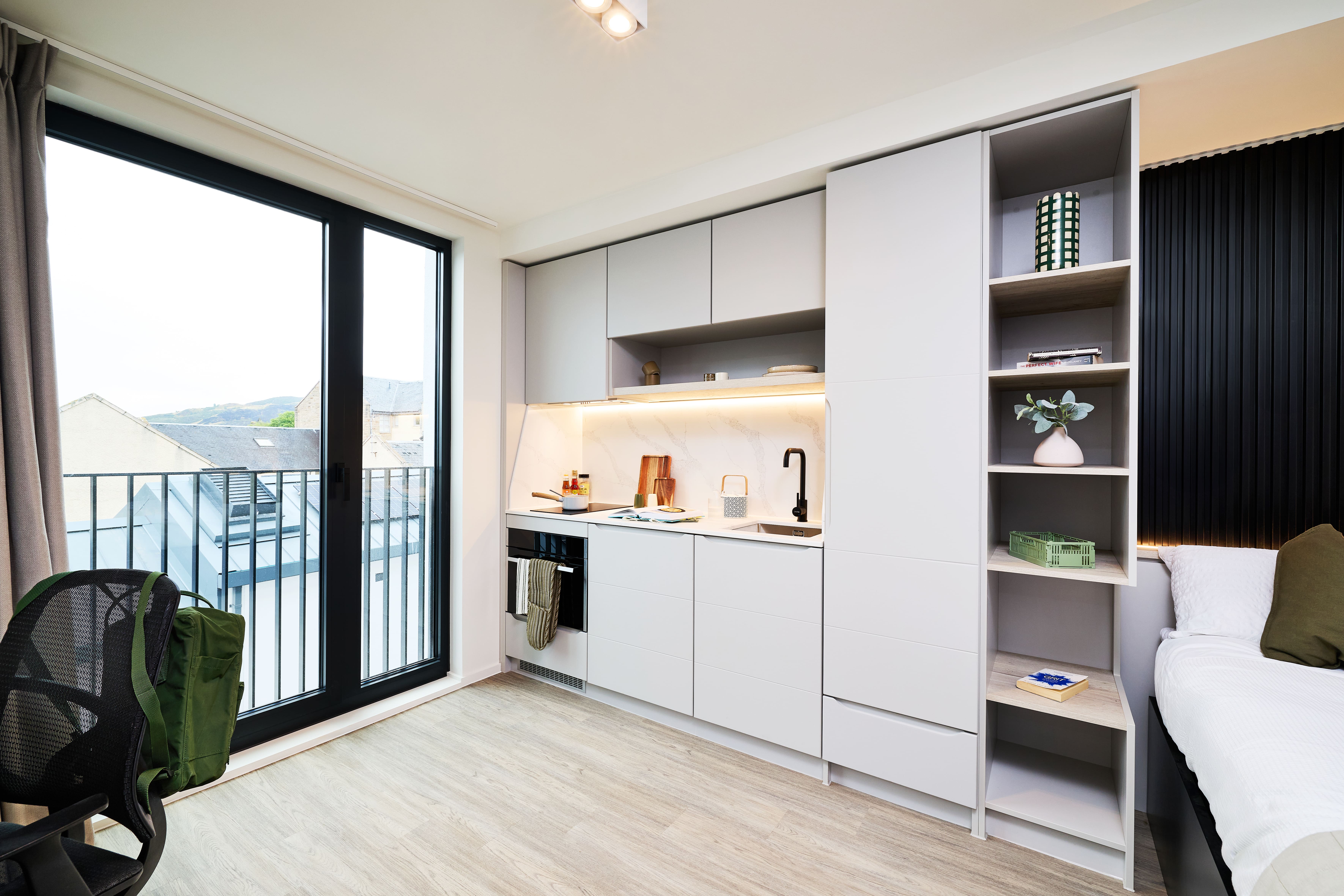 Modern kitchenette with light gray cabinets, integrated appliances, and open shelving next to a sliding glass door and a bed.
