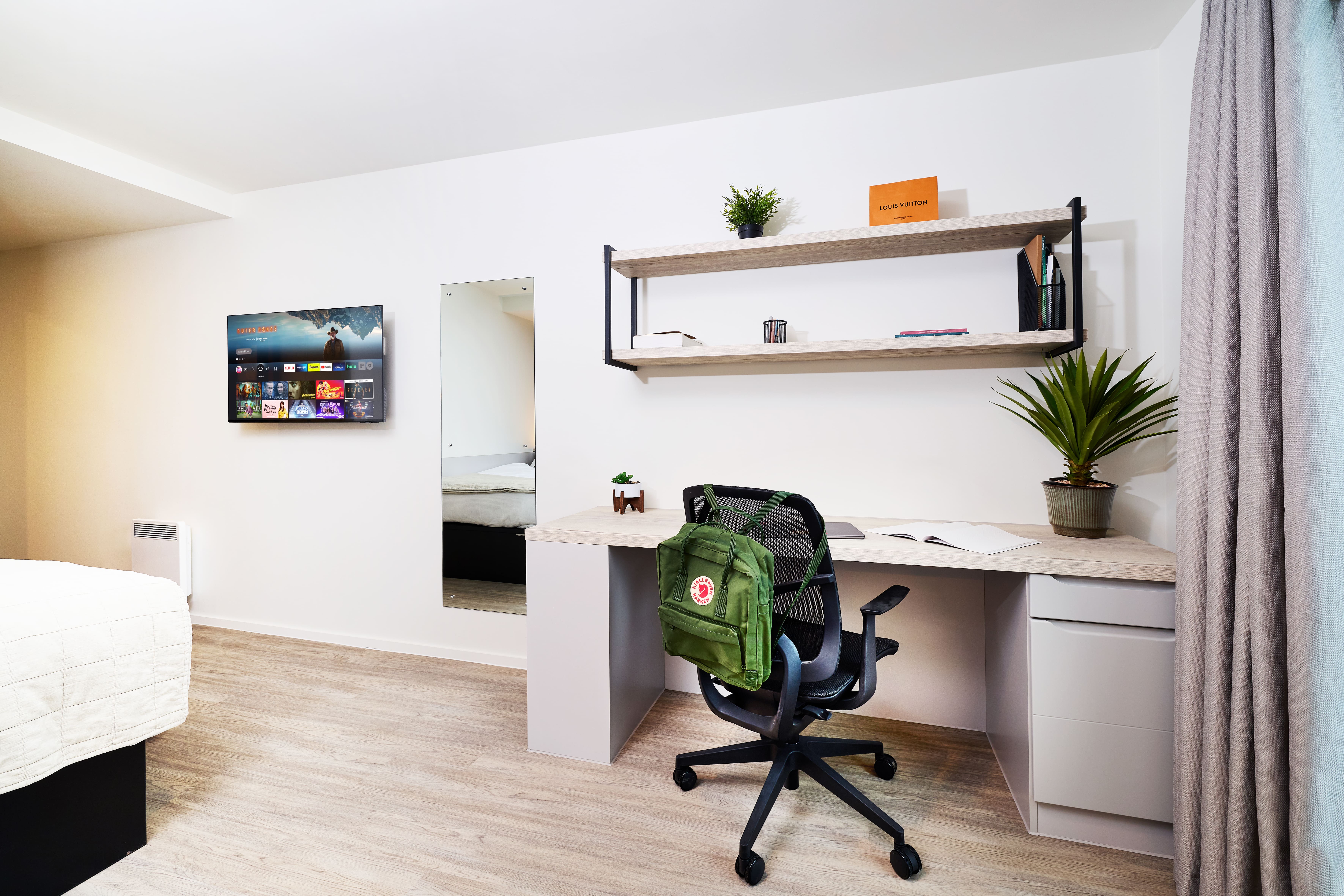 A desk with an office chair and a green backpack, positioned below shelves and a wall-mounted television in a bedroom.