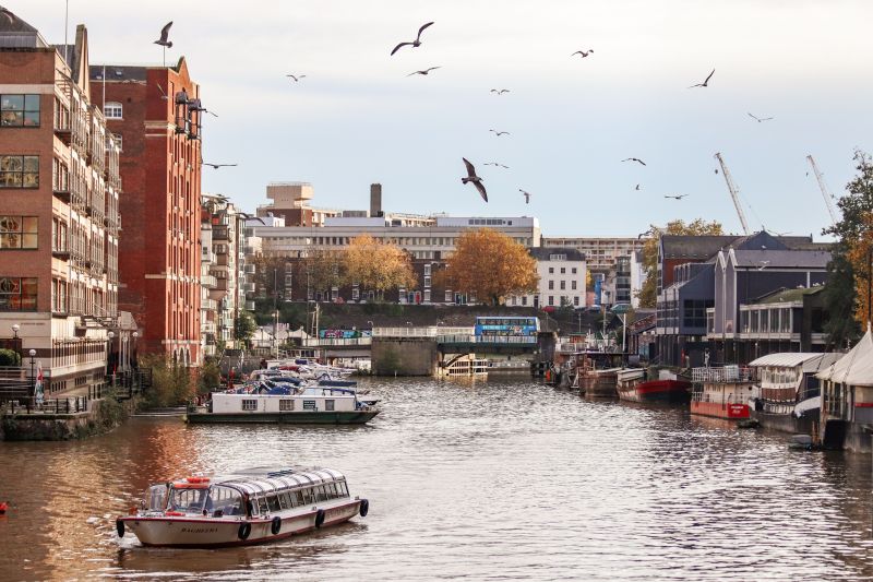 A tour boat travels down a Birmingham city river lined with buildings, moored houseboats, and several birds flying overhead, a popular route for student travel.