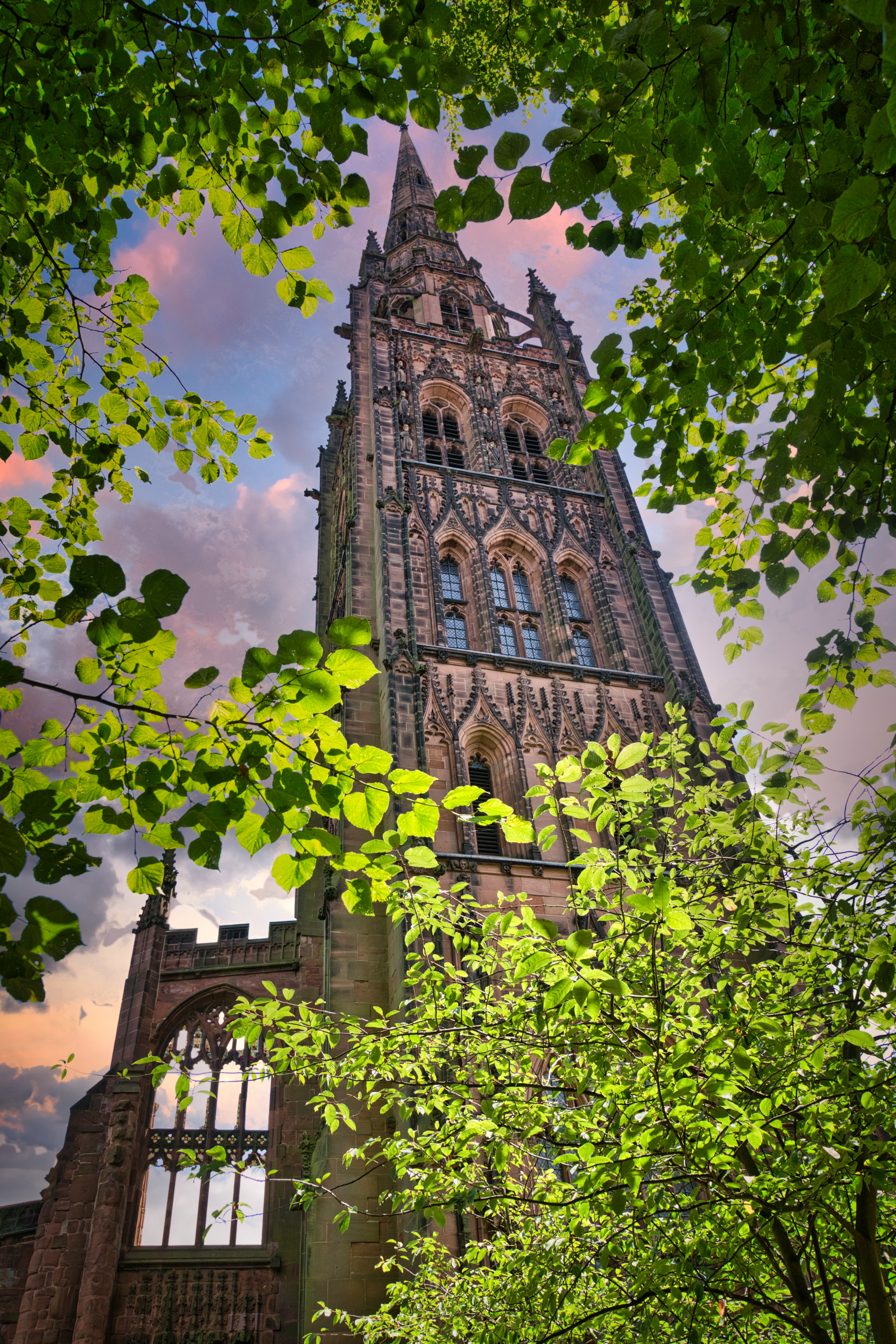 A tall, ornate stone church spire framed by green tree branches against a cloudy, sunset-colored sky, located near student accommodation in Coventry.