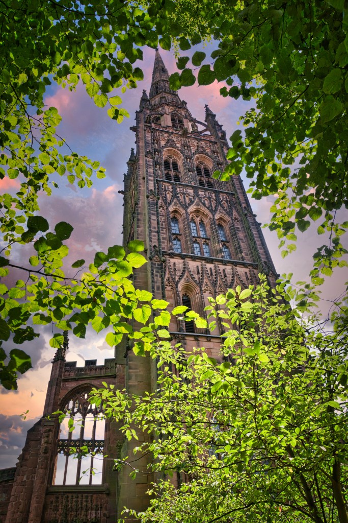A tall, ornate stone church spire framed by green tree branches against a cloudy, sunset-colored sky, located near student accommodation in Coventry.