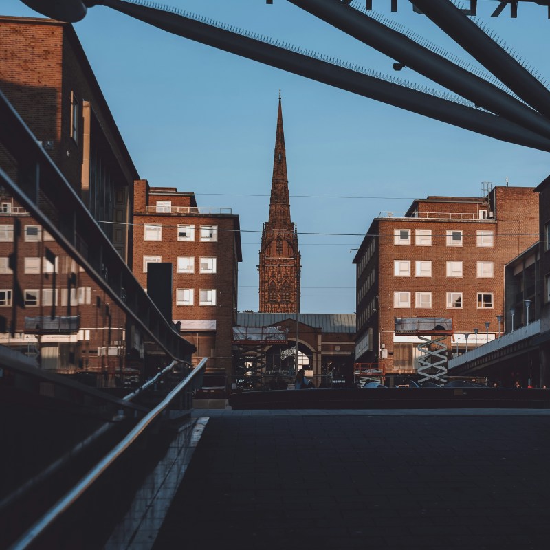 A view between modern student accommodation in Coventry toward a tall, slender church spire, framed by an overhead structure.