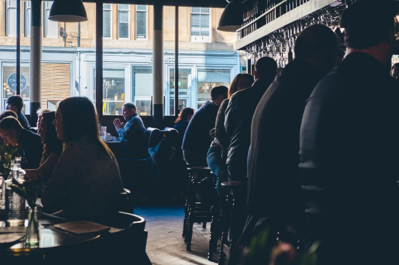 Patrons sit at tables and a bar inside a dimly lit restaurant, with large windows looking out onto a bright city street near student accommodation in Glasgow.