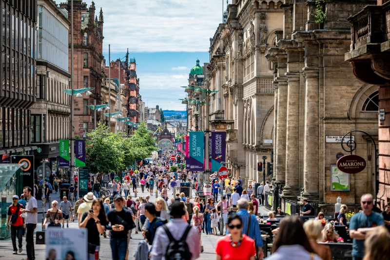 A crowded pedestrian street in Glasgow, near popular student accommodation, featuring stone buildings, outdoor cafe seating, and banners promoting the 2018 European Championships.