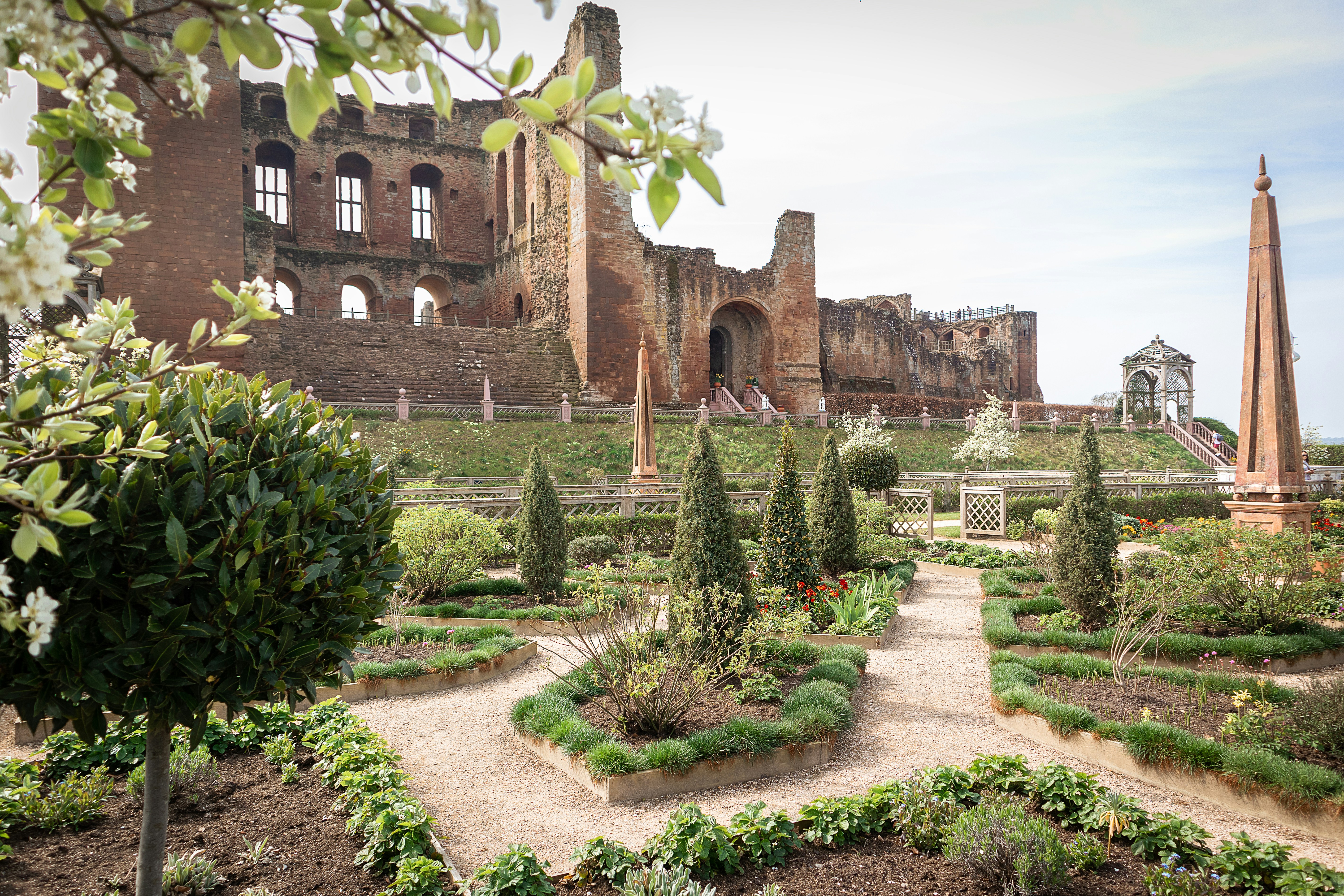 A manicured garden with shaped shrubs and gravel paths in front of the stone ruins of Kenilworth Castle, a popular destination for student travel in Birmingham.