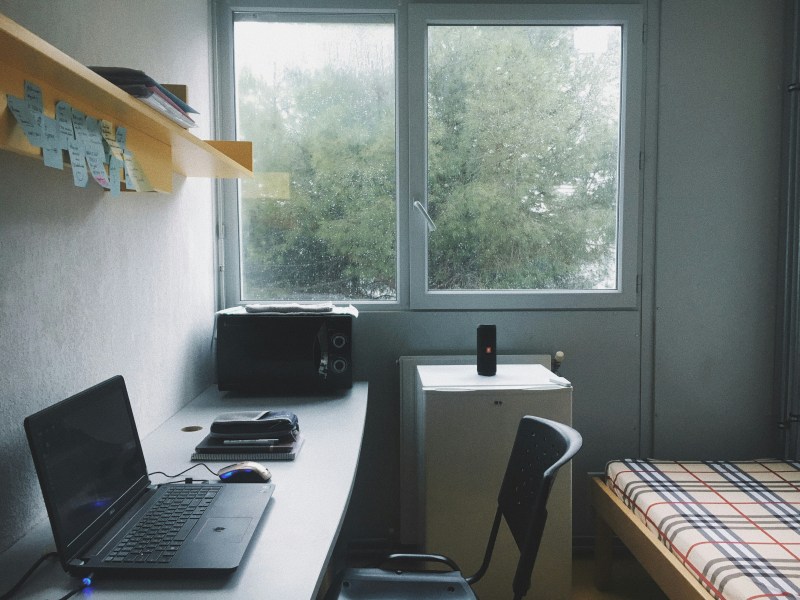 A compact student housing room with a desk, laptop, and mouse by a window overlooking trees, featuring a single bed and a small refrigerator with a speaker.