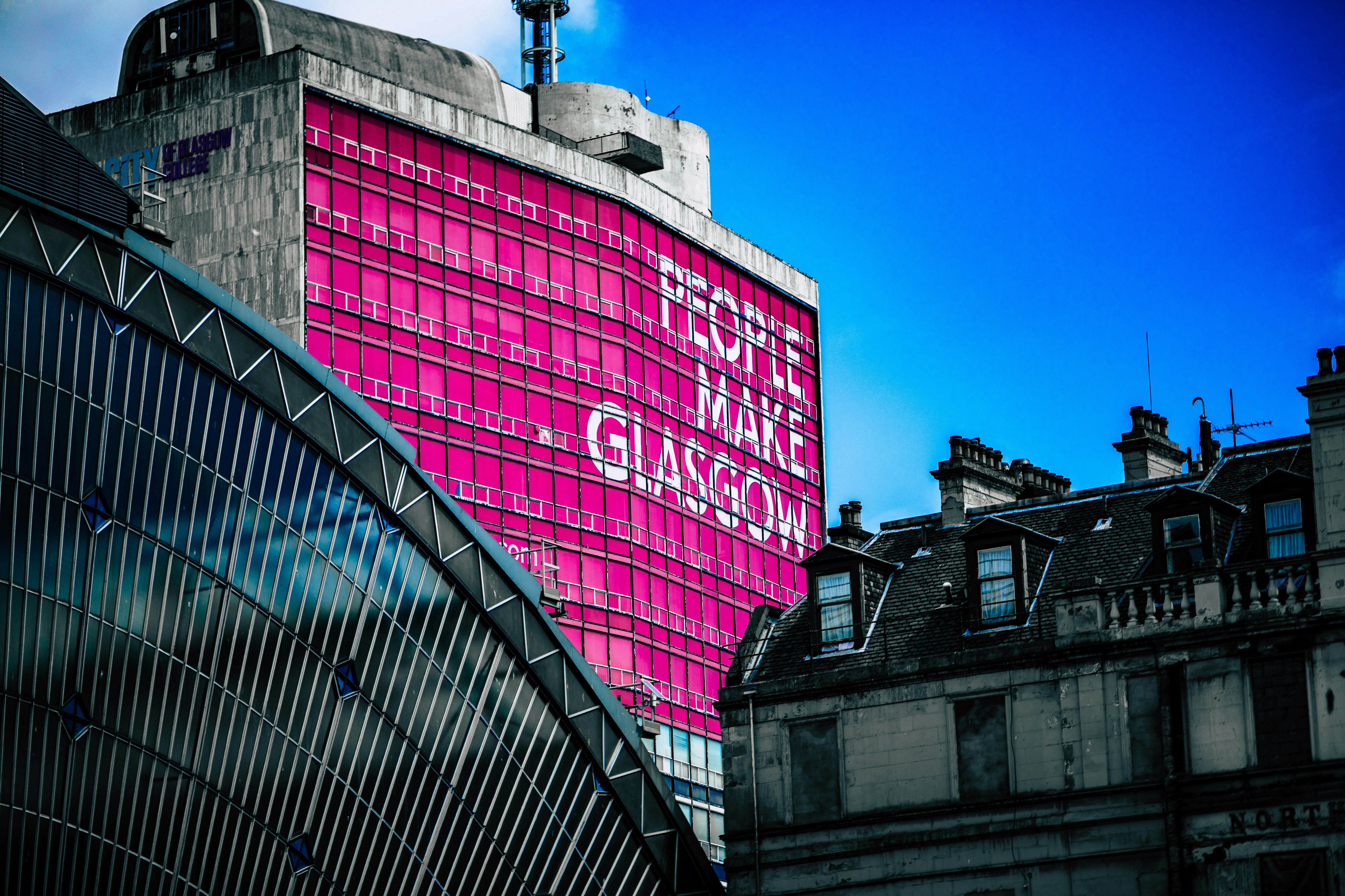 A large building with a bright pink facade featuring the text “PEOPLE MAKE GLASGOW” serves as modern student accommodation in Glasgow, situated next to a domed structure and historic stone buildings.