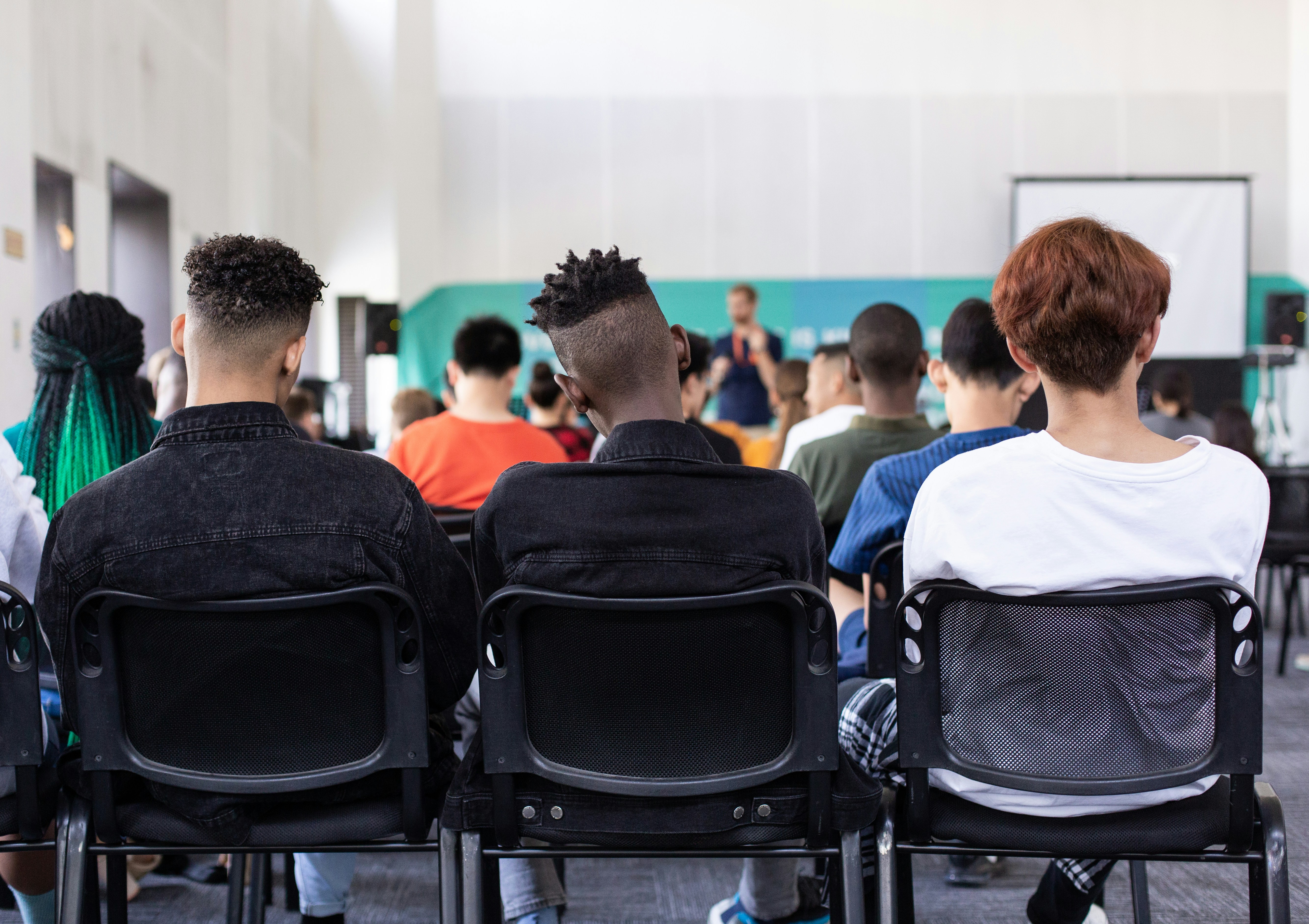 A group of people sitting in chairs in a student housing common area, viewed from behind, facing a speaker at the front of the room.