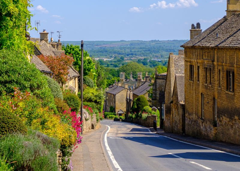 A road descends between traditional stone houses and lush, colorful flower gardens toward a rolling green valley, a perfect scene for student travel day trips from Birmingham.